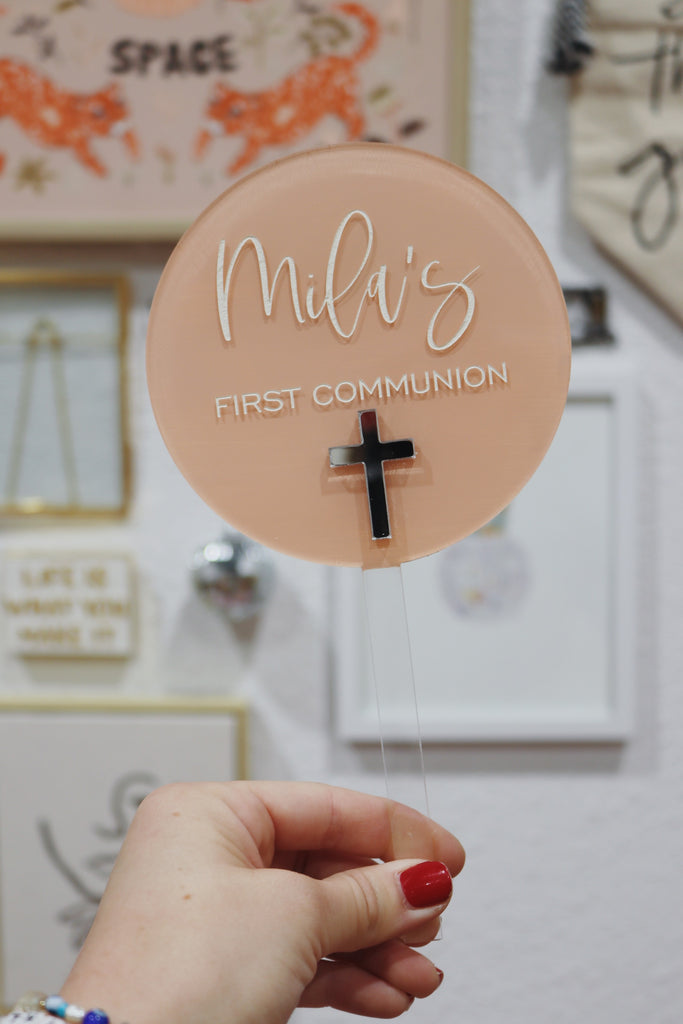 Round acrylic ornament with 'Mila's First Communion' text and cross, held by a hand.