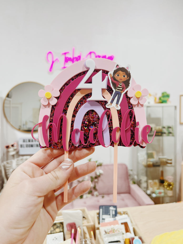 Hand holding a personalized birthday cake topper with a character and decorative elements.